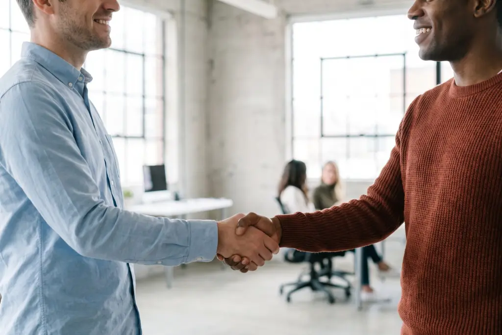Due persone si stringono la mano in un ufficio, con colleghi al tavolo sullo sfondo